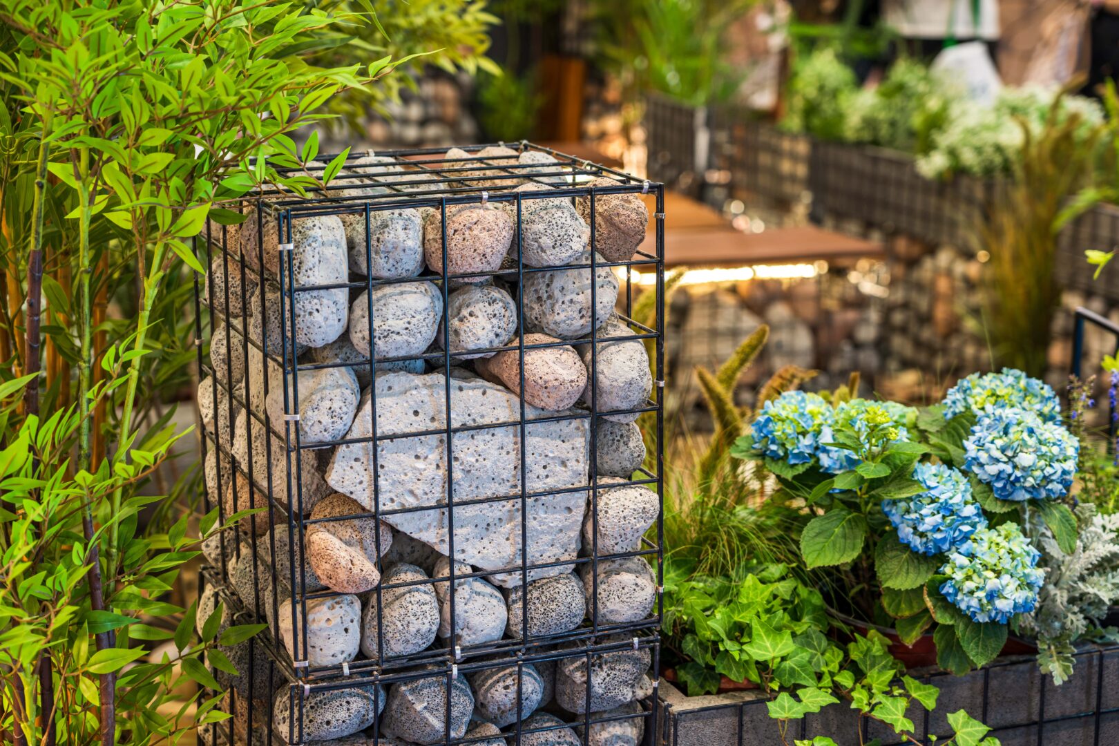 Eine mit großen Steinen gefüllte Gabione steht neben Grünpflanzen und blauen Hortensien in einem Garten im Freien.