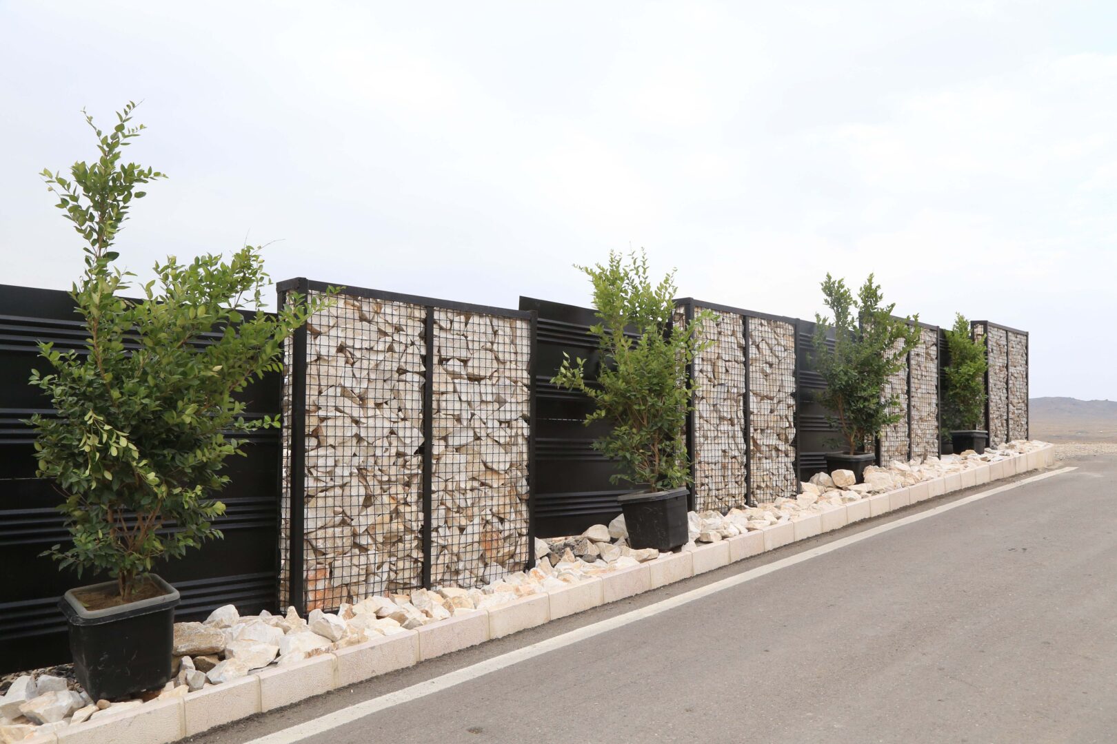 Eine Straßensperre aus schwarzen Metallplatten und mit Steinen gefüllten Drahtkäfigen mit jungen Bäumen, die in Containern entlang des Gehwegs gepflanzt wurden.